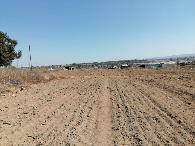A wide-angle view of a vacant residential plot featuring cleared earth, perimeter fencing, and distant neighborhood structures under a clear sky.