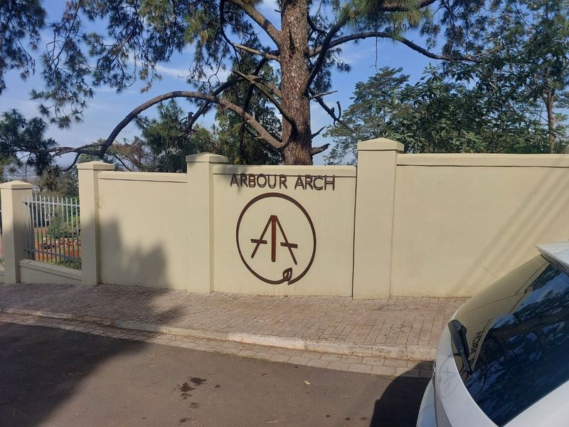 The entrance facade of Arbour Arch featuring a solid boundary wall with signage, paved sidewalk, and established trees.
