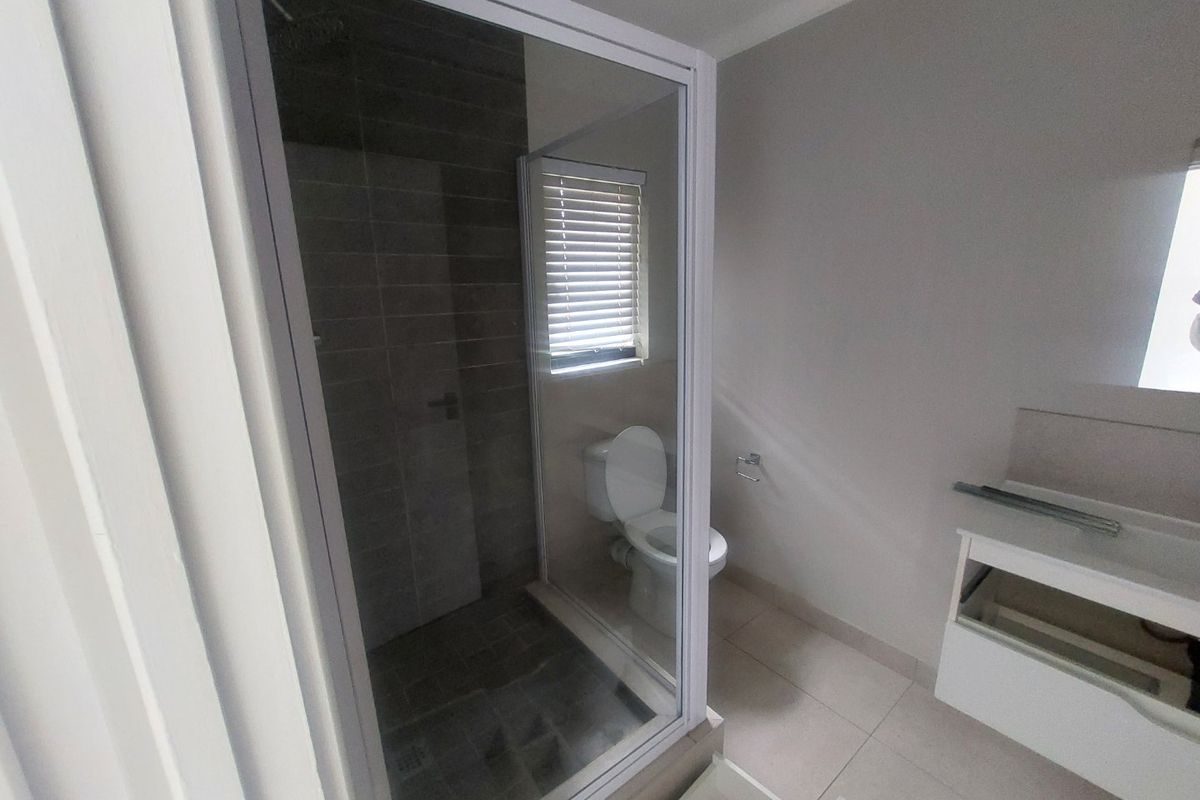 A functional bathroom featuring a glass-enclosed shower, toilet, and a vanity unit with a missing drawer front, finished with neutral floor and wall tiles.