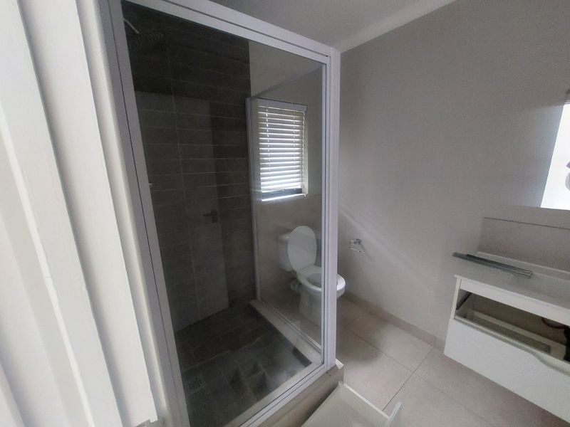 A functional bathroom featuring a glass-enclosed shower, toilet, and a vanity unit with a missing drawer front, finished with neutral floor and wall tiles.
