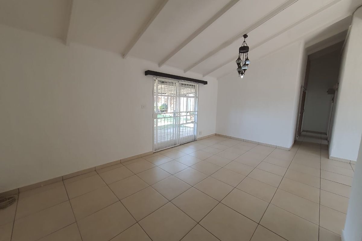 A bright, empty living room featuring tiled flooring, exposed beam ceilings, and a sliding glass door with a security gate leading to the exterior.
