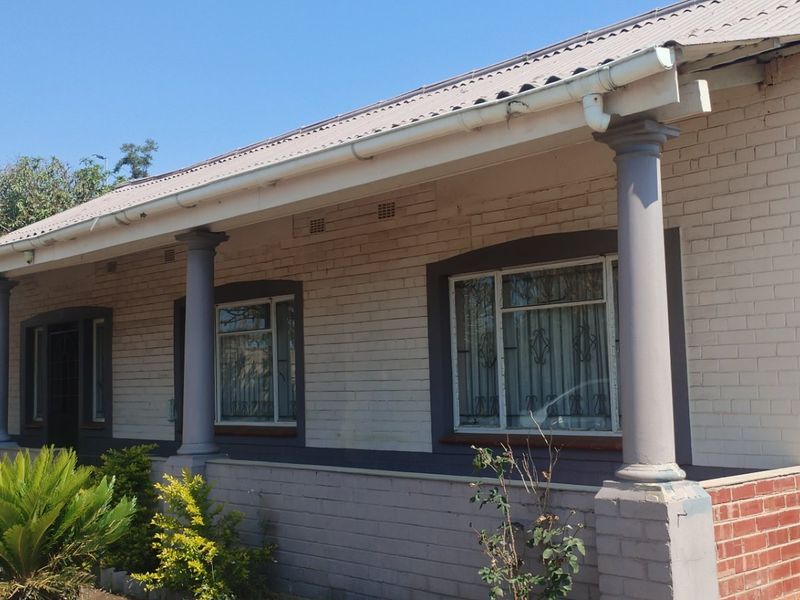 Front elevation of a brick house featuring a covered veranda with concrete columns, corrugated metal roofing, and security bars on the windows.