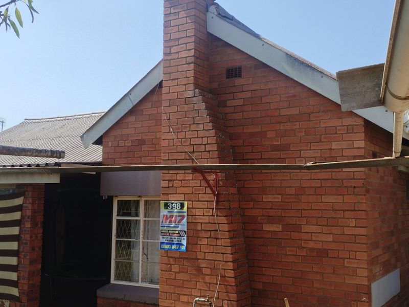 Rear view of a brick house featuring a prominent chimney, corrugated roofing, and security bars on the windows.