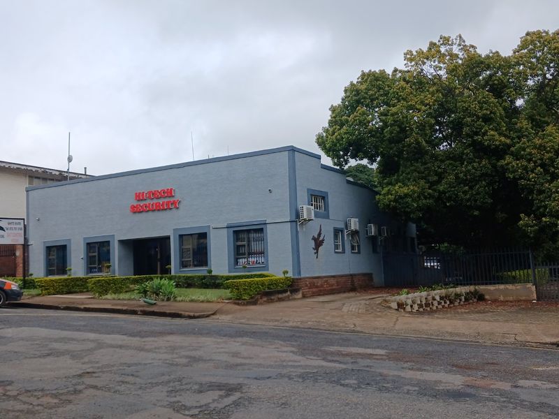 Commercial property exterior featuring a blue facade, security signage, wall-mounted air conditioning units, and established garden beds along the street frontage.