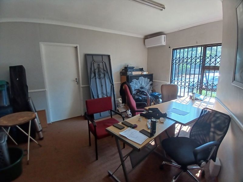 A functional office space featuring carpeted flooring, a wall-mounted air conditioning unit, and a window secured with a metal security gate.