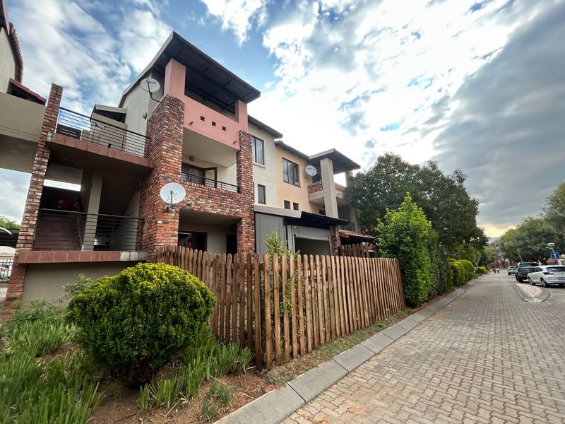 Exterior view of a multi-story apartment complex featuring brick accents, private balconies, and a wooden picket fence along the paved street.
