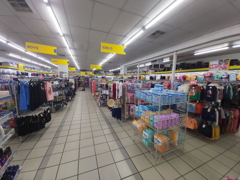 A spacious commercial retail interior featuring tiled flooring, suspended ceiling grid with fluorescent lighting, and organized clothing display aisles.