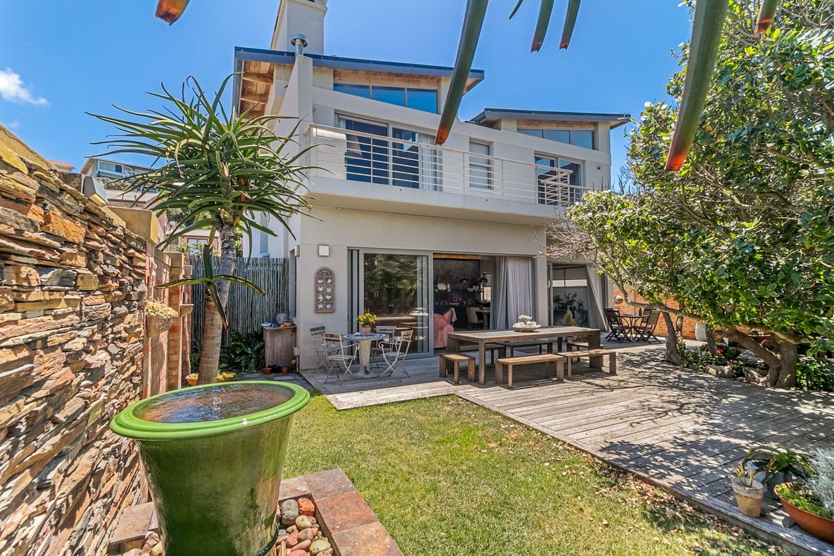 Rear view of the property featuring a timber deck, landscaped garden, stone retaining wall, and a balcony accessible from the upper level.