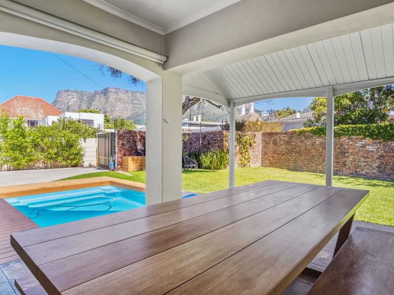 A covered outdoor patio overlooking a swimming pool, lawn, and brick-walled garden with mountain views in the background.