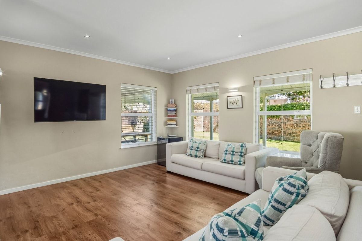A bright and well-maintained living room featuring laminate flooring, recessed downlighting, and large windows with venetian blinds overlooking the garden.