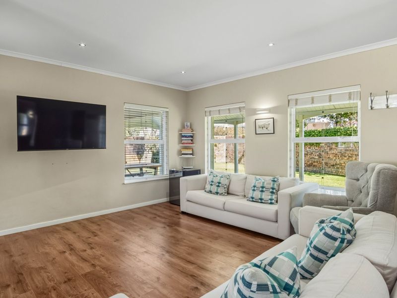 A bright and well-maintained living room featuring laminate flooring, recessed downlighting, and large windows with venetian blinds overlooking the garden.