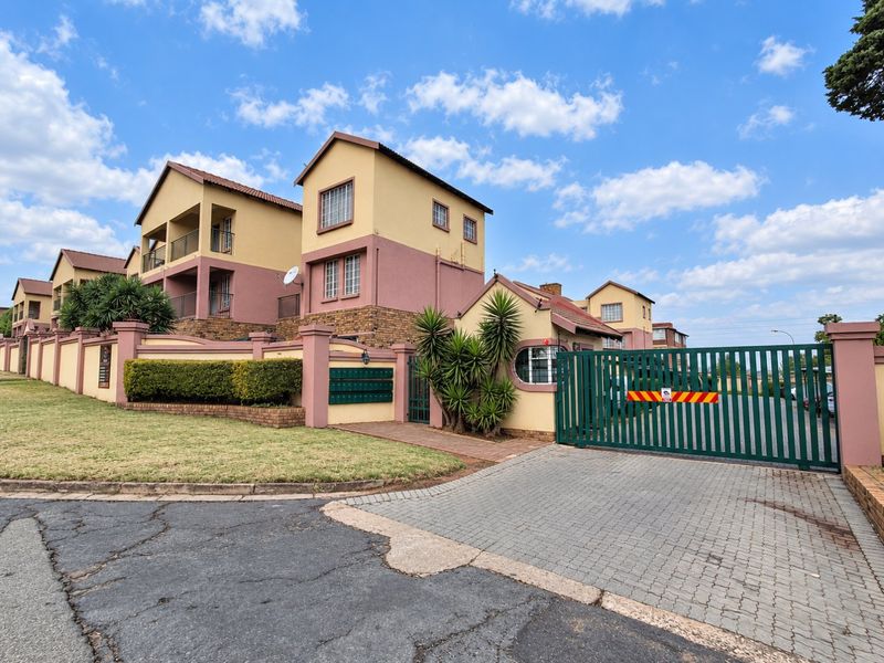 Front view of a townhouse complex featuring a secure gated entrance, paved driveway, and multi-story residential units with balconies.