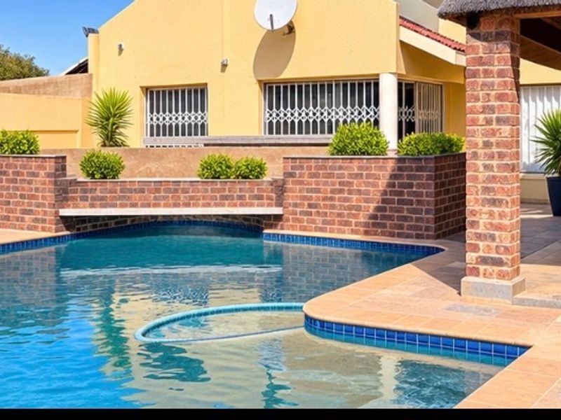 A well-maintained outdoor pool area featuring a tiled patio, brick retaining wall, and a covered entertainment space with brick pillars.