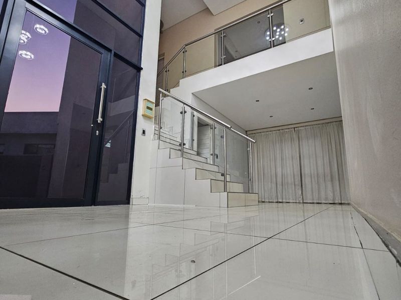 Spacious double-volume entrance hall featuring modern tiled flooring, a sleek glass balustrade staircase, and contemporary pendant lighting.