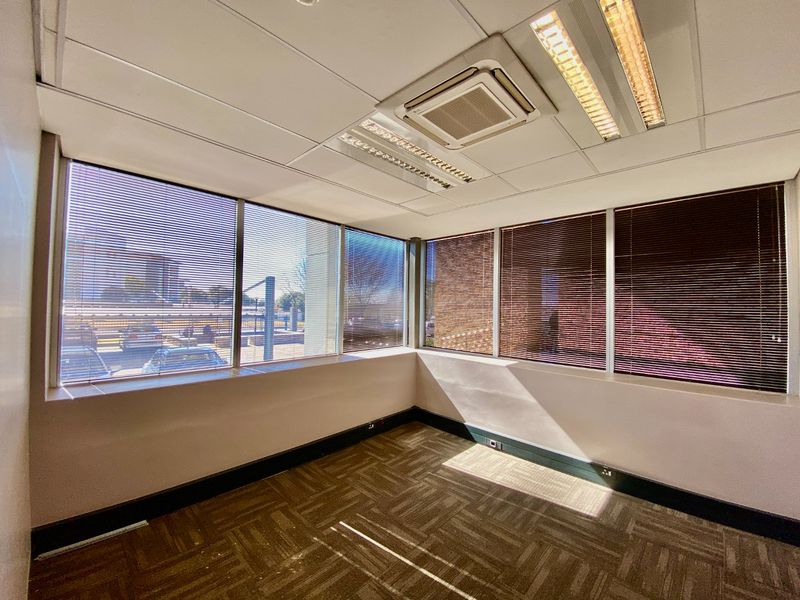 A bright, empty corner office space featuring large windows with venetian blinds, carpeted flooring, and a suspended ceiling with integrated air conditioning.
