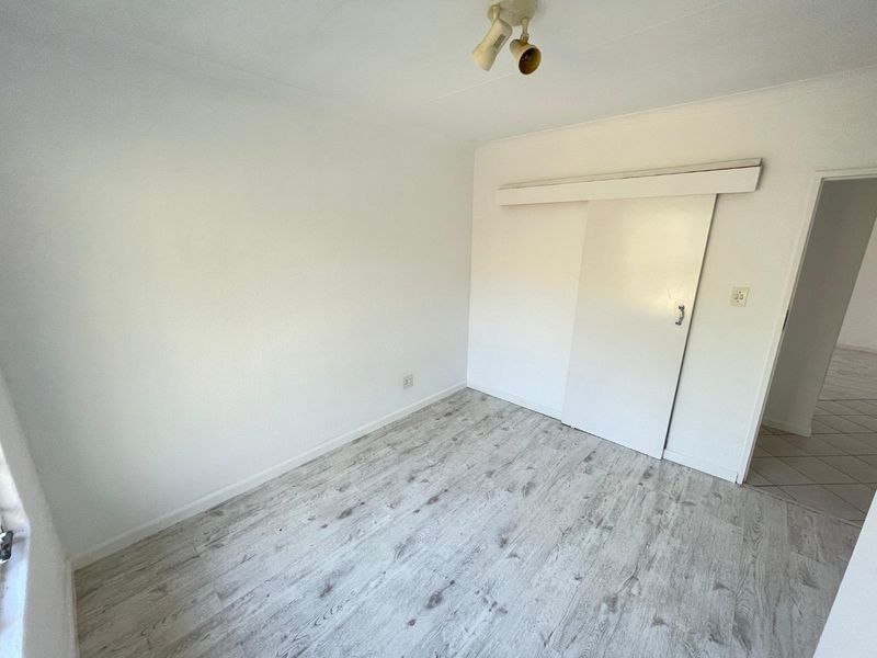 An empty bedroom featuring light-toned laminate flooring, a sliding door, and a ceiling-mounted spotlight fixture, offering a clean and neutral space.