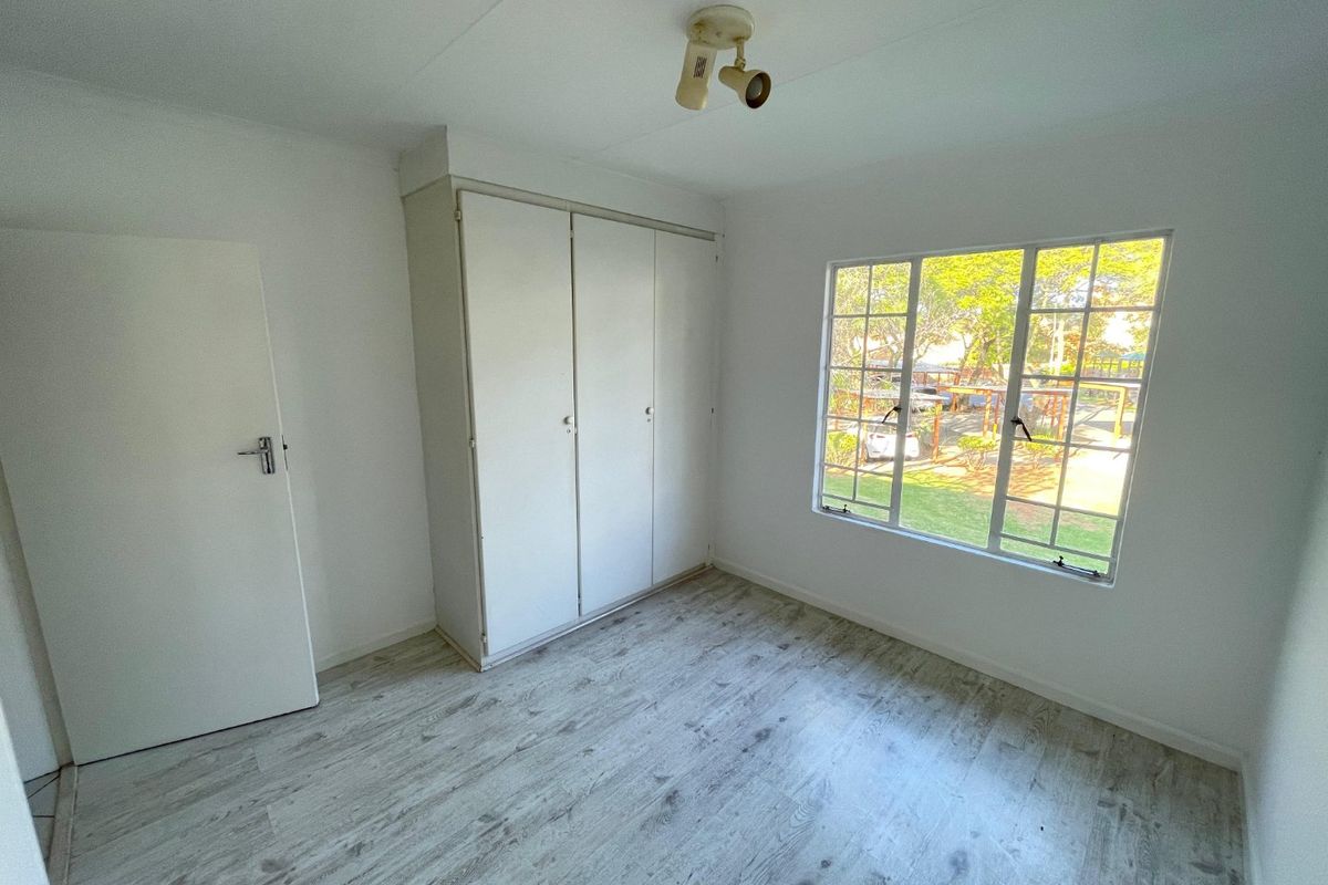 A bright, empty bedroom featuring light-toned laminate flooring, a built-in wardrobe, and a window with security bars overlooking the garden.