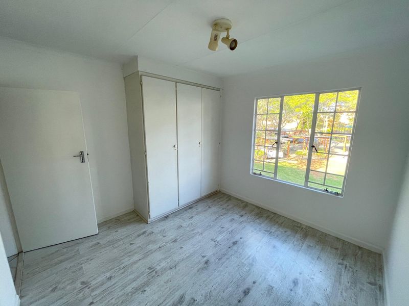 A bright, empty bedroom featuring light-toned laminate flooring, a built-in wardrobe, and a window with security bars overlooking the garden.