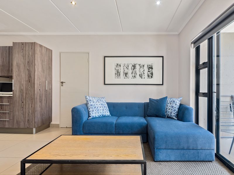 Modern open-plan living area featuring tiled floors, recessed lighting, and a compact integrated kitchen with wood-grain cabinetry, leading to a private balcony.