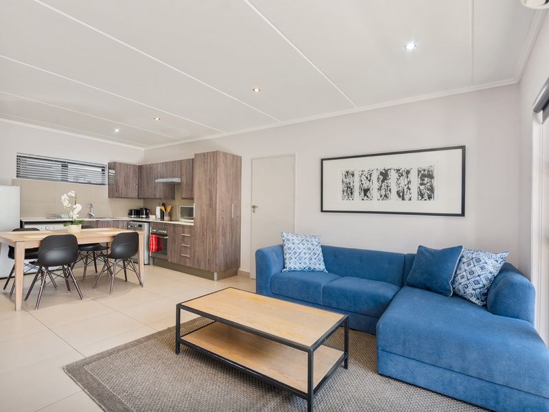 A bright open-plan living and kitchen area featuring modern wood-finish cabinetry, integrated appliances, tiled flooring, and recessed lighting throughout.