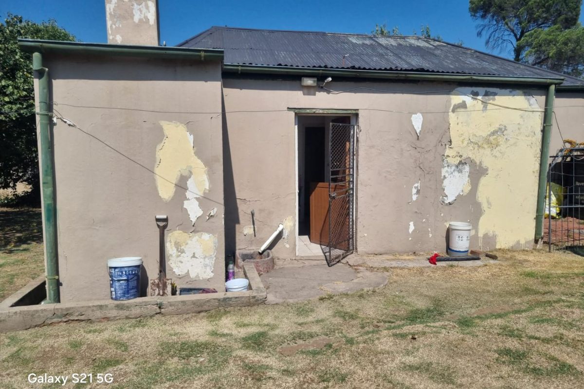 Rear exterior view of the property showing significant paint peeling and plaster damage on the walls, with a corrugated metal roof and security gate.