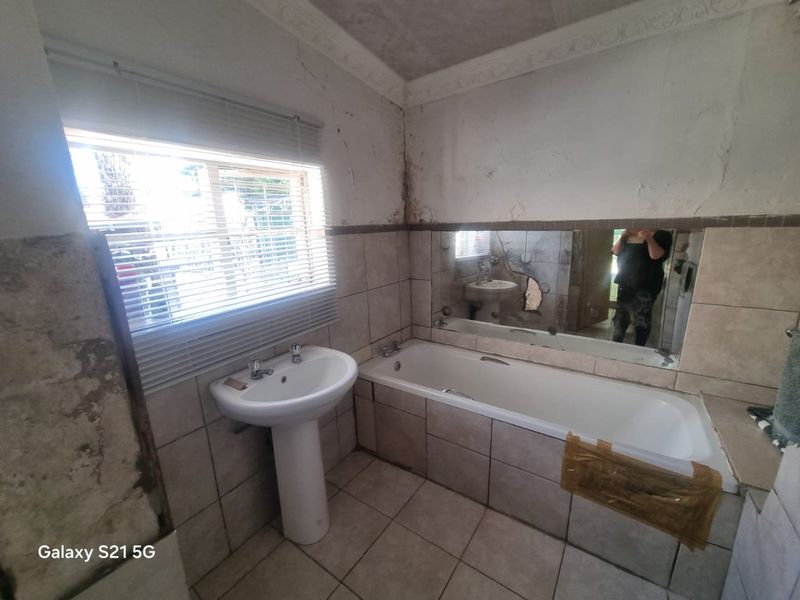 A bathroom in need of renovation, featuring a pedestal basin, built-in bathtub, and tiled finishes with visible wall damage and a cracked mirror.