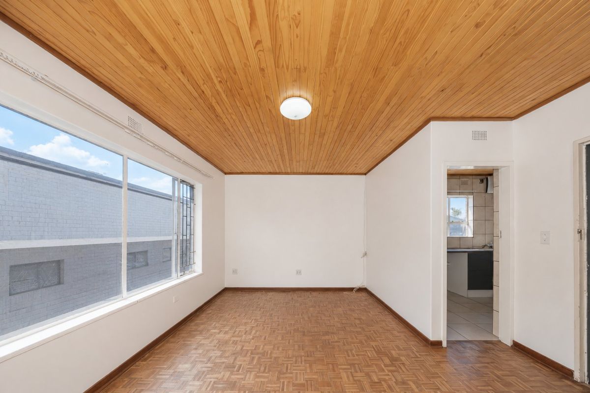 An empty living room featuring parquet flooring and a timber-clad ceiling, with large windows providing natural light and a view into the adjacent kitchen area.
