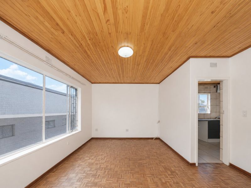 An empty living room featuring parquet flooring and a timber-clad ceiling, with large windows providing natural light and a view into the adjacent kitchen area.
