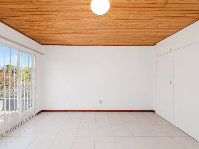 A bright, empty bedroom featuring tiled flooring, built-in cupboards, a timber ceiling, and a sliding door leading to a balcony with security gate.