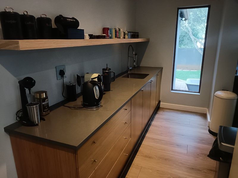 A modern scullery or coffee station featuring sleek wood-finish cabinetry, stone countertops, an undermount sink, and a floating shelf with natural light.