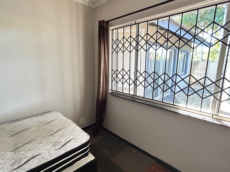 A simple bedroom featuring a large window with a fitted security gate, neutral walls, and carpeted flooring.