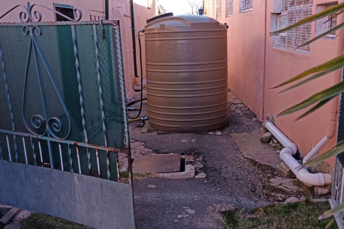 Rear exterior utility area featuring a large water storage tank, wrought iron gate, and exposed external plumbing along the building wall.