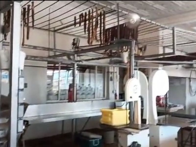 Commercial food processing or butchery utility space featuring industrial-grade stainless steel work surfaces, overhead hanging racks, and heavy-duty band saw equipment.