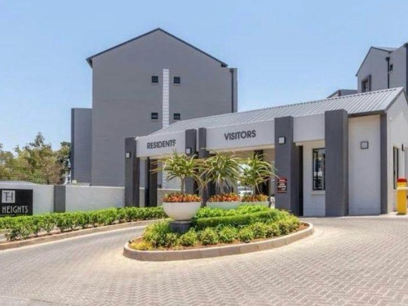 The main entrance and security gatehouse of The Heights apartment complex, featuring a paved driveway, boom gate access, and landscaped traffic island.