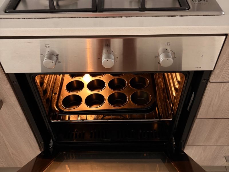 A close-up view of a modern stainless steel gas hob and built-in oven integrated into a stone countertop and light wood-finish cabinetry.
