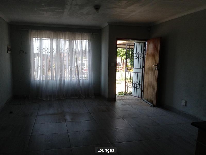 An empty living room featuring tiled flooring, a security gate at the entrance, and a timber door, requiring cosmetic updates and improved lighting.