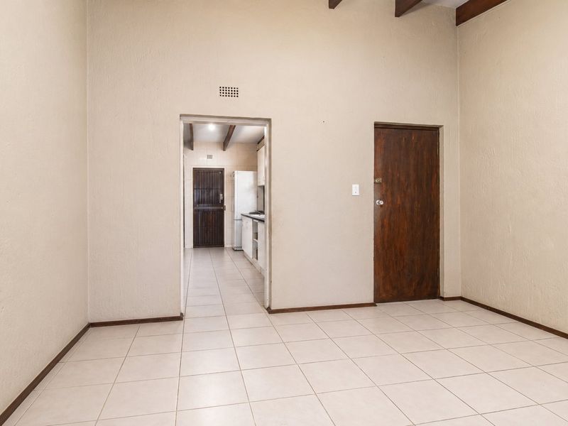 An empty living area featuring neutral tiled flooring and exposed timber ceiling beams, with a doorway leading through to the kitchen area.