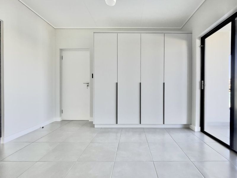 A bright, modern bedroom featuring neutral tiled flooring, ample built-in cupboard storage, and a sliding glass door providing access to the exterior.