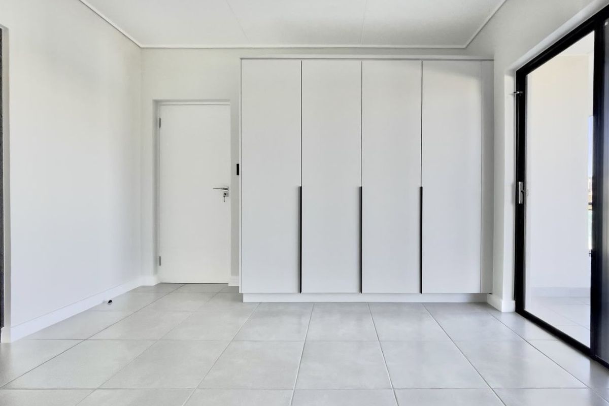 A bright, modern bedroom featuring neutral tiled flooring, ample built-in cupboard storage, and a sliding glass door providing access to the exterior.