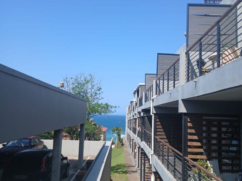 Exterior view of the apartment complex featuring tiered balconies with metal railings, a covered parking area, and a pathway leading toward the ocean.