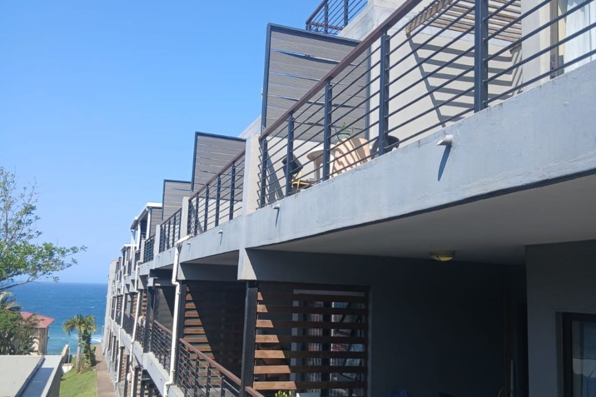 A side view of a multi-story apartment complex featuring tiered balconies with metal railings and timber privacy screens, overlooking a coastal landscape.