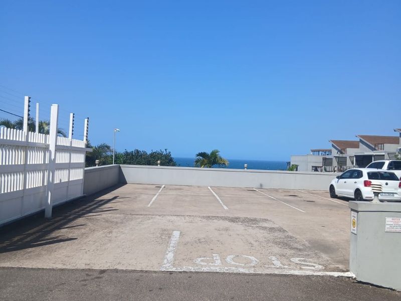 An open-air parking area featuring marked bays, electric fencing, and a concrete boundary wall with a distant ocean view.