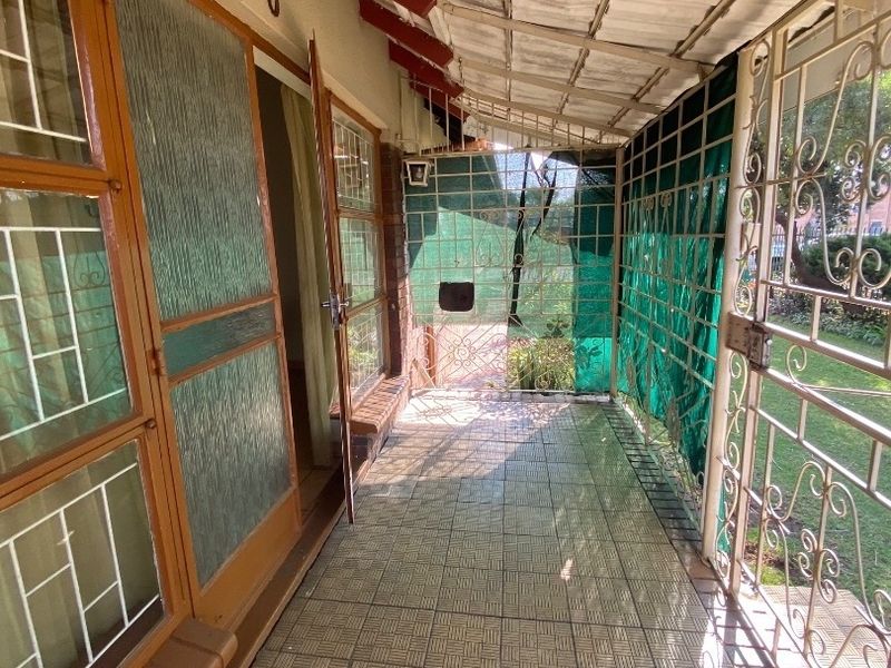 A covered outdoor patio area featuring tiled flooring, security gates, and a metal roof structure, providing direct access to the garden.