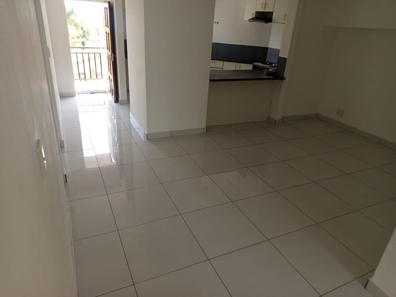 An open-plan living area featuring neutral ceramic floor tiles, leading to a kitchen with built-in cabinetry and a breakfast bar, with balcony access.