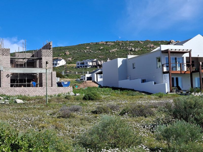 A view of a residential plot featuring an ongoing construction project alongside an existing finished home in a coastal landscape setting.