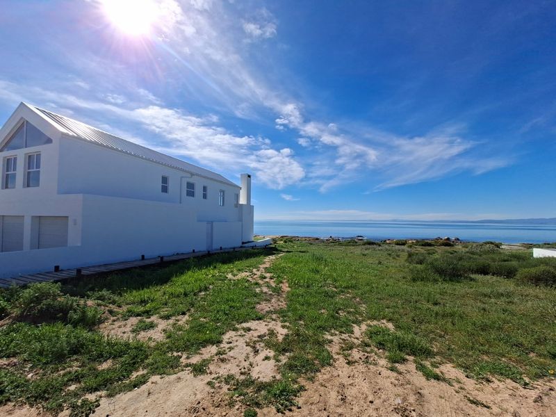 A rear view of a modern white coastal residence featuring a metal roof, chimney, and a timber boardwalk leading toward the ocean.