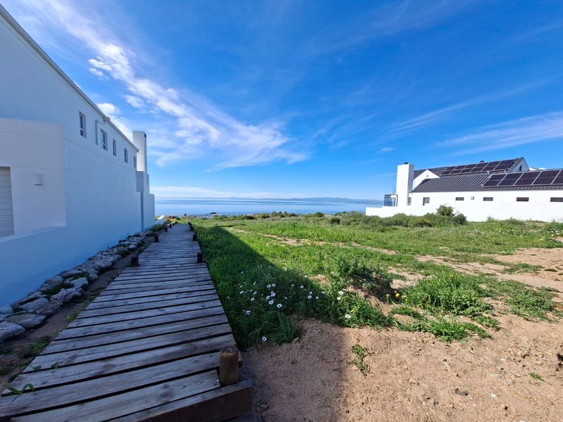 A scenic rear exterior view featuring a wooden boardwalk leading towards the coastline, with modern residential buildings and solar panels visible.