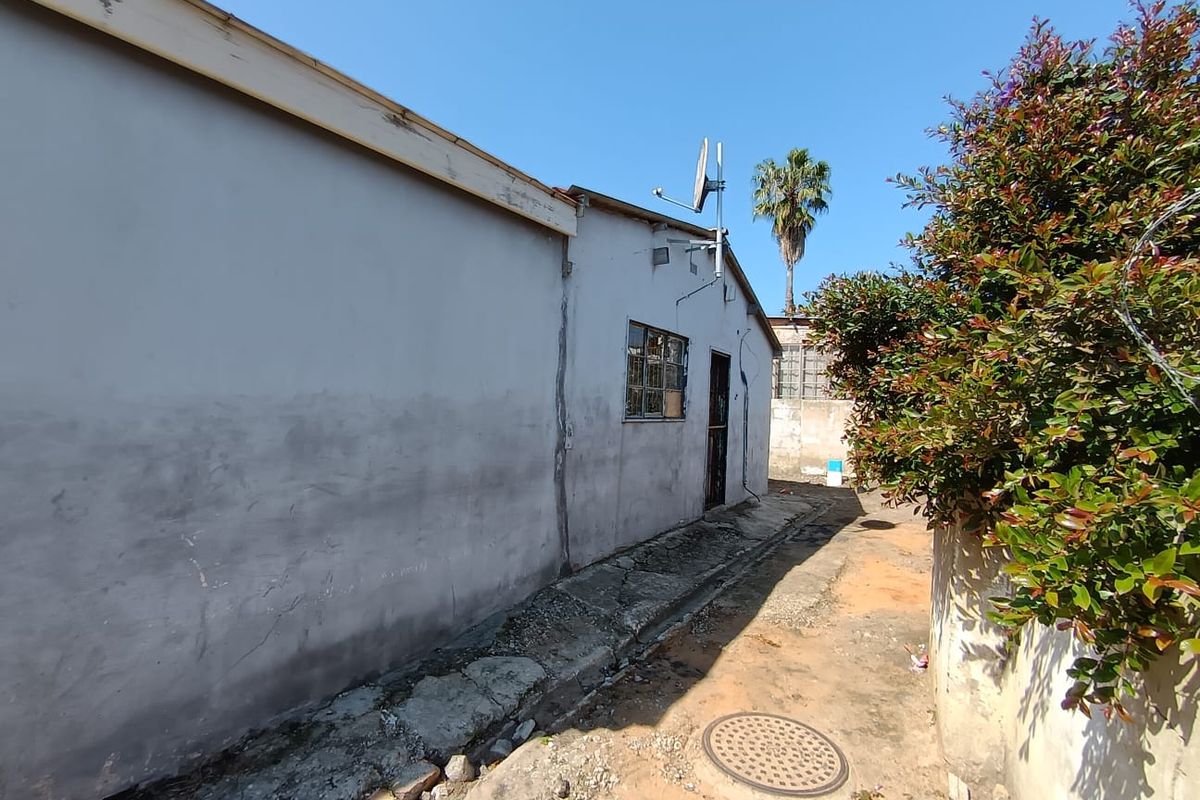 A view of the rear exterior wall showing signs of weathering, cracked plaster, and a concrete pathway leading along the side of the house.