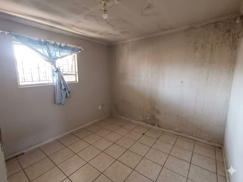 An empty bedroom featuring tiled flooring and security bars on the window, currently showing significant signs of damp and water damage on walls and ceiling.