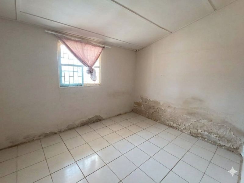 An empty bedroom featuring tiled flooring and a window with security bars, currently requiring significant wall repairs and cosmetic refurbishment.